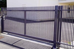 A modern black aluminum fence gate with vertical bars stands in front of a white building, casting long shadows on the ground. This image highlights the sleek design and durability of aluminum gates, showcasing one of the many aluminum fence gate colors available for residential or commercial properties.