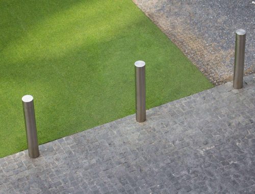 Three sturdy metal posts are installed in a paved driveway, evenly spaced and aligned, with a grassy area and gravel in the background. This image illustrates how to install driveway gate posts, showcasing proper placement and secure installation techniques for durability and functionality.