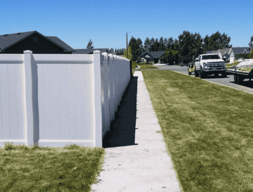 newly installed white vinyl privacy fence runs alongside a clean sidewalk in a suburban neighborhood under a bright blue sky. In the background, work trucks and fencing materials are visible. This image highlights expert tips for maintaining your fence in Ammon's climate, ensuring durability and longevity in varying weather conditions.