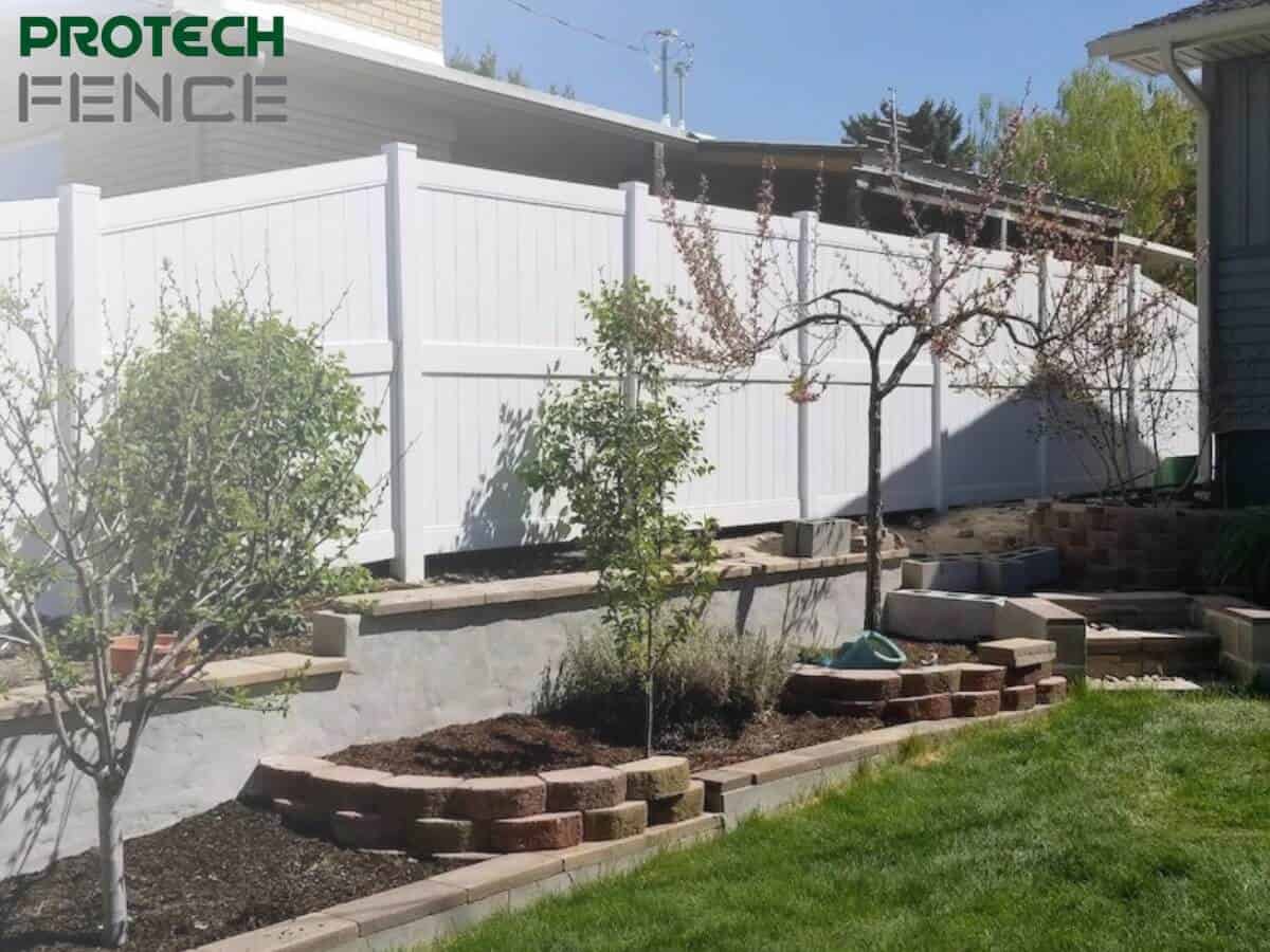 A serene backyard featuring Protech Fence’s 10 ft vinyl fence gate, characterized by its bright white color providing privacy and security. The gate complements a well-maintained garden with brick-edged flower beds, young trees, and a multi-level stone landscaping element, enhancing the home’s outdoor living space. 