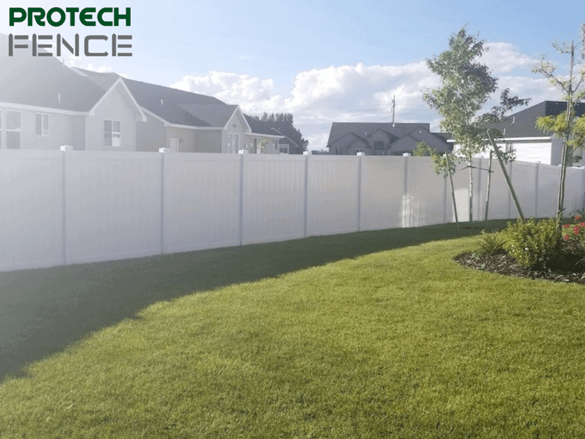 A neatly maintained backyard showcasing the clean lines of a white vinyl privacy fence, illustrating an example of the white vinyl fence cost per foot for residential properties. The fence encloses a lush green lawn with a small, circular garden bed featuring vibrant red and yellow flowers, complemented by a young tree and an ornamental garden cart. The fence provides privacy and a serene backdrop to the charming outdoor space.