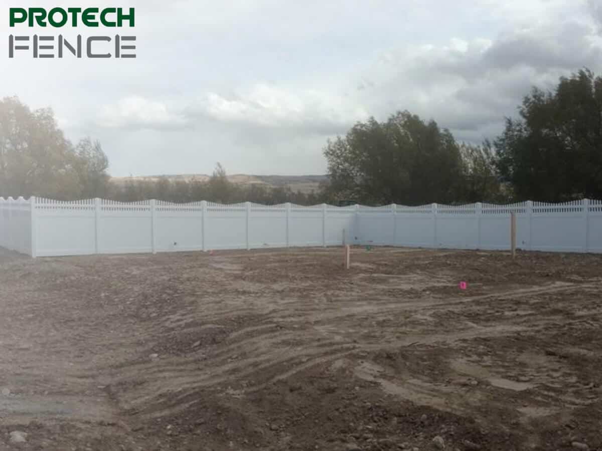 A lengthy 12 foot white vinyl fence runs across a developing landscape. The scene is overcast, with a rough, muddy construction site in the foreground. Scattered trees stand behind the fence under a grey sky, suggesting a large, open area in the midst of development. 