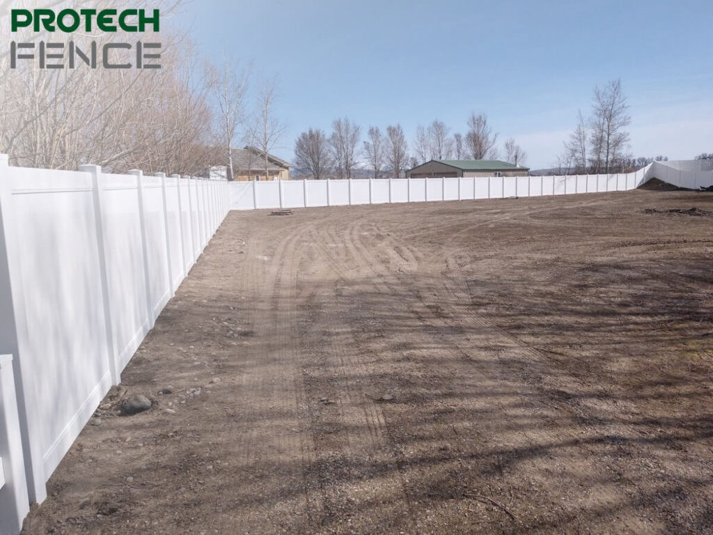 A panoramic view of a property bordered by a pristine 8 foot vinyl fence gate. The vinyl fencing is stark white and extends in a long, unbroken line across the landscape, indicating a vast, secure perimeter. The ground in the foreground is bare soil with track marks, suggesting recent installation or maintenance work, with a residential area and trees visible in the background under a clear sky.