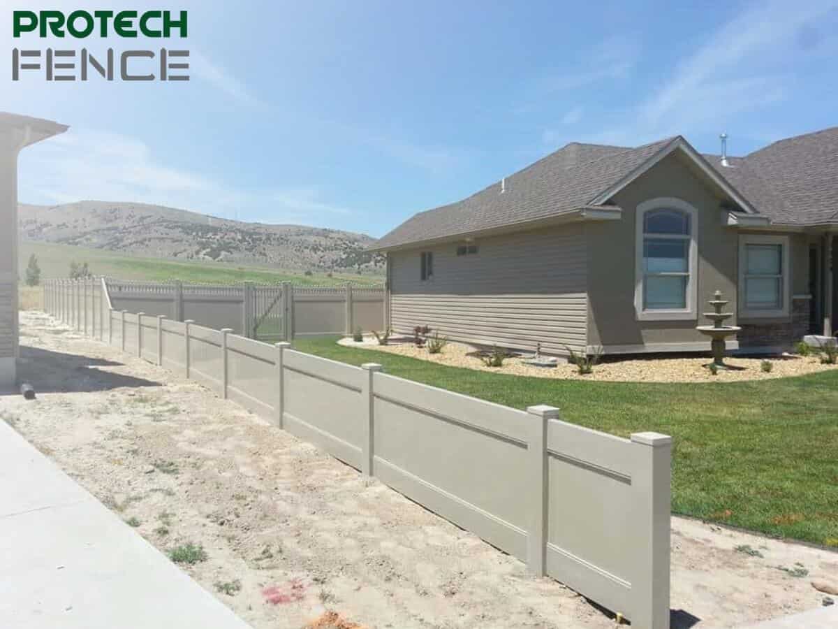 A vinyl fence installation pocatello extends around the perimeter of a residential yard, with the Protech Fence logo in the top left, showcasing a beige fence against a backdrop of a house and hills, providing a secure and private boundary.