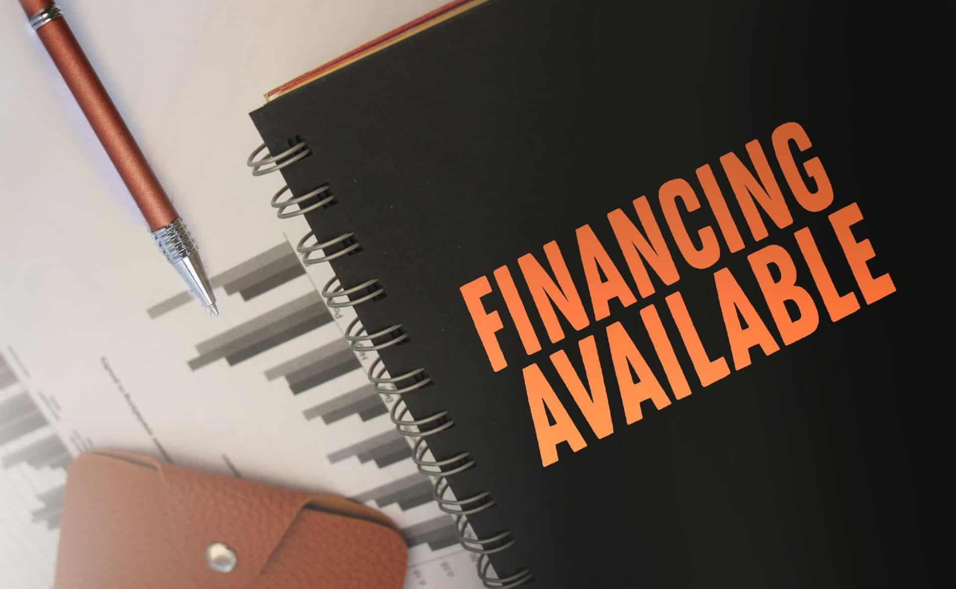 An open notebook with "Financing Available" written in bold orange text, emphasizing a fence payment plan near me. A pen and financial charts are visible in the background, suggesting financial planning and budgeting options.