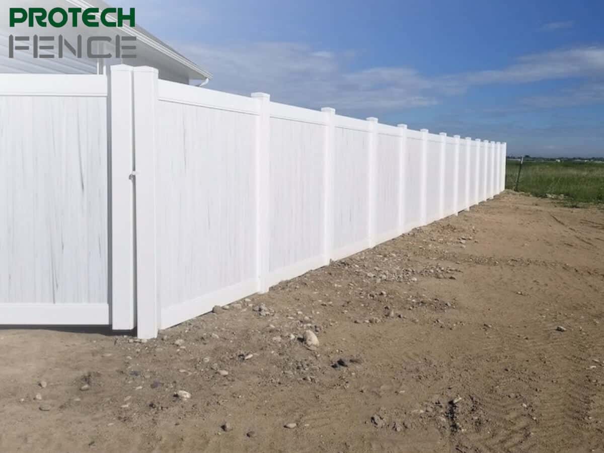  This image shows a 12 foot tall white vinyl fence. The fence runs along a rough dirt path with sparse grassy terrain in the background under a clear blue sky. The fence’s clean lines and solid panels offer a stark contrast to the rugged ground alongside it.